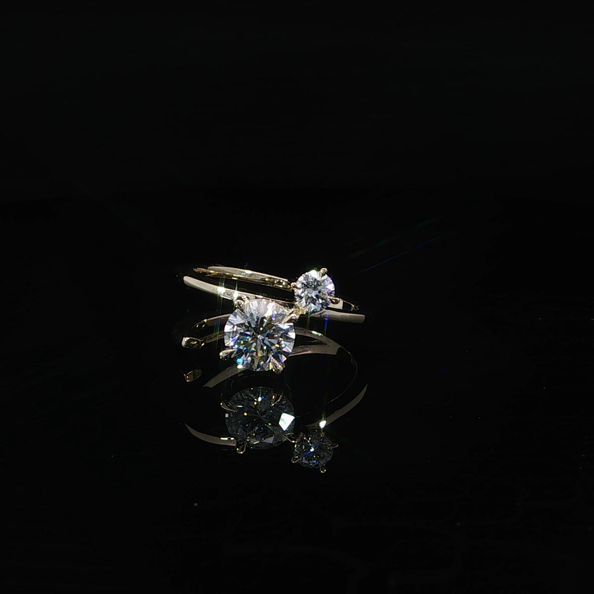 Two round diamonds arranged diagonally on an open yellow gold band, reflected on a black surface.