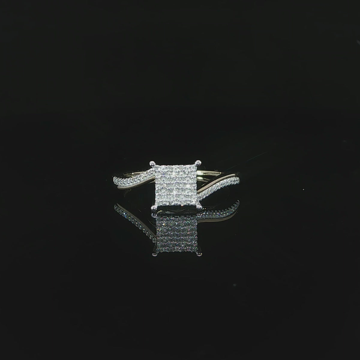 Video of square diamond cluster and curved yellow gold shoulders reflected on a glossy black surface.