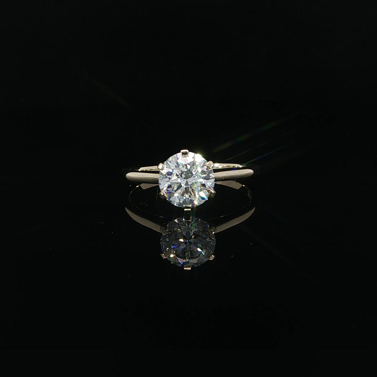 Front view of a six-prong round diamond solitaire reflected on a glossy black surface.